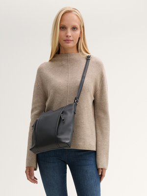 Svart texturerad axelväska med två blixtlåsfickor och ett justerbart band. Bärs av en person i en beige tröja och blå jeans.
