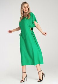 Grünes Midikleid mit kurzen Ärmeln, Seitenausschnitten und verstellbaren Bindebändern in der Taille. Glatter Stoff mit fließender Silhouette. Schwarze Absätze.