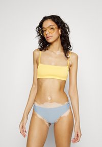 Top de bralette amarelo canelado com alças finas, combinado com um fundo de bikini texturizado azul claro. A modelo tem cabelo encaracolado e óculos transparentes.