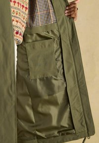 Jachetă verde măslin cu interior căptușit în carouri și un buzunar cu etichetă. Materialul pare neted și izolat.