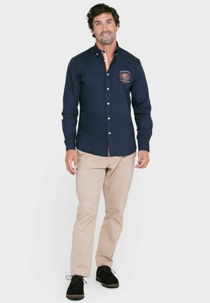 Camisa de botón de manga larga azul marino con un logo en el pecho, cuello de color contrastante, combinada con pantalones beige y zapatos negros.