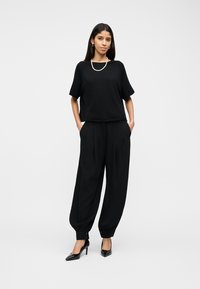 Femme debout, les mains dans les poches, portant un haut noir à manches courtes, un pantalon noir ample avec des poignets à boutons, des talons noirs et un collier de perles.