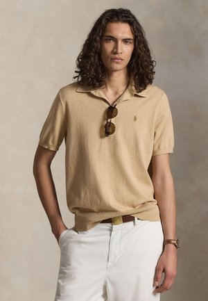 Junger Mann mit langen lockigen Haaren, trägt ein beiges Poloshirt, weiße Hose, Sonnenbrille am Kragen hängend und eine braune Uhr am linken Handgelenk.