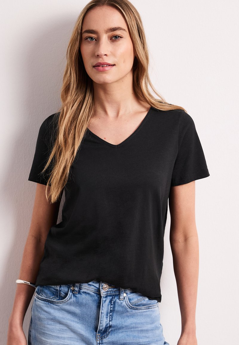 Femme aux longs cheveux blonds portant un T-shirt noir à col en V et un jean bleu clair, debout contre un mur blanc.