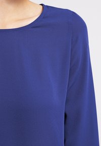 Lähikuva henkilöstä, joka käyttää yksiväristä pitkähihaista kuninkaansinistä yläosaa pyöreällä pääntiellä vaaleaa taustaa vasten.