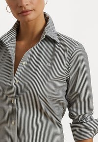 Mustavalkoraidallinen napitettava paita, jossa on kaulus, pieni brodeerattu logo ja käärityt hihat. Sileä puuvillakangas.