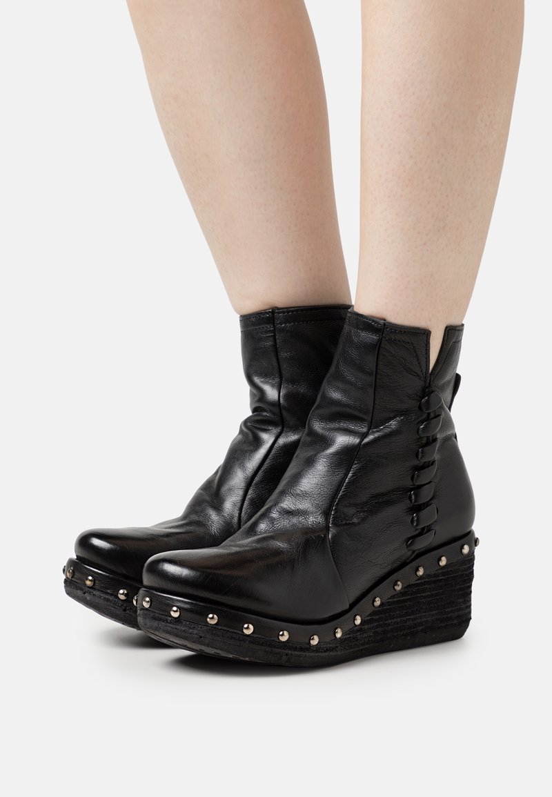 Botas de tobillo de cuero negro con un tacón en cuña en capas, que presentan acentos de tachuelas y detalles de cordones laterales. Textura suave, diseño elegante.