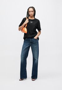 Μαύρο t-shirt με λογότυπο, μπλε τζιν φαρδιάς γραμμής και πορτοκαλί τσάντα χειρός. Η εμφάνιση ολοκληρώνεται με μαύρα σανδάλια με λουράκια.