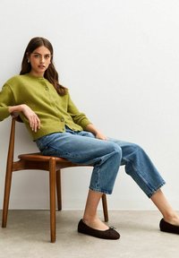 Groene gebreide cardigan met ronde hals, knoopsluiting; lichtblauwe wijde jeanstroeven; bruine ballerina's; zittend op een houten stoel tegen een lichte muur.