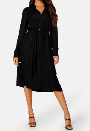 Kobieta ubrana w długą czarną sukienkę midi na guziki z wiązaniem w talii oraz czarne sandały na obcasie, pozująca na jednolitym białym tle.