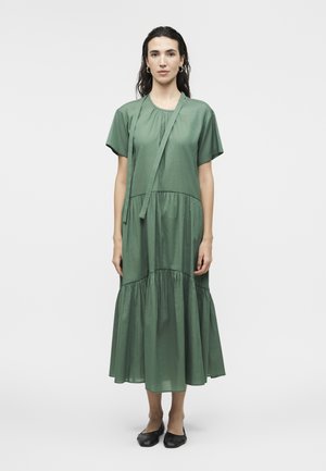 Γυναίκα που στέκεται φορώντας ένα άνετο πράσινο μίντι φόρεμα με κοντά μανίκια και μαύρα επίπεδα παπούτσια μπροστά σε απλό φωτεινό φόντο.