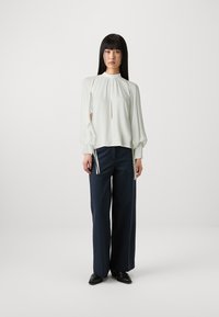 Blusa verde chiaro con un colletto alto, maniche lunghe a sbuffo e dettagli a taglio. Abbinata a pantaloni larghi blu navy e scarpe nere.