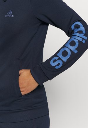 Sweat-shirt Adidas bleu marine avec la main gauche dans la poche, petit logo sur la poitrine et grand texte bleu Adidas courant le long de la manche droite.