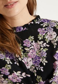 Sort blomsterbluse med rynket hals. Designet inkluderer lilla og grønne blomster med fine detaljer mod en struktureret baggrund.