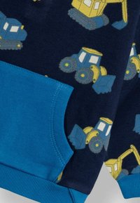 Centrāla jūras svīteris ar dzeltenu un zilu būvniecības transportlīdzekļu rakstu; tam ir vienkrāsains zils ribots aproces un kabatas akcents.