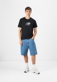 T-shirt noir avec logo argenté, associé à un short en denim bleu clair. Porté avec des chaussettes blanches et des baskets grises. Tenue simple et décontractée.