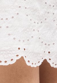 Λευκό ύφασμα με μικρές οπές και κυματιστό τελείωμα που δείχνει κεντητά λουλουδένια σχέδια και μικρές κυκλικές περικοπές πάνω σε φόντο χρώματος δέρματος.