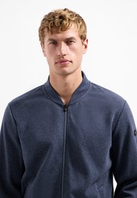 Marineblauwe ritsjack met een gestructureerde stof, opstaande kraag en zijzakken. Beschikt over een klein logo op de linker mouw.