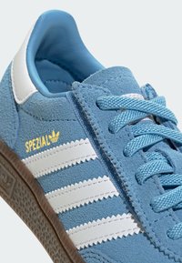 Modré semišové tenisky se třemi bílými pruhy, texturovaným svrškem a modrými tkaničkami. Mají hnědou gumovou podrážku a nápis 'SPEZIAL' ve zlaté barvě.
