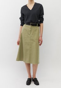 Donkergrijze v-hals trui gecombineerd met een lichtgroene midi-jurk met twee voorzakken en een zwarte riem. Zwarte loafers maken de outfit compleet.