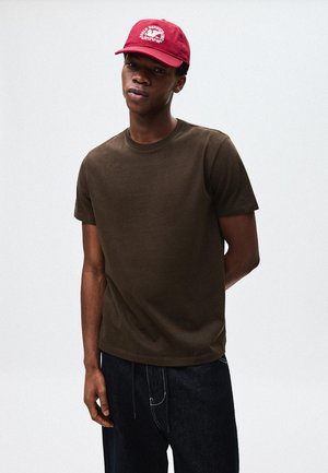 Smeđa pamučna majica s okruglim izrezom i kratkim rukavima, nošena s tamnoplavim trapericama. Crvena baseball kapa s detaljem logotipa.