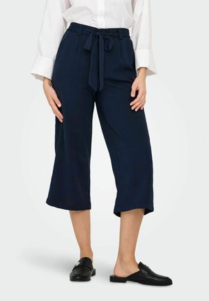 Femeie purtând pantaloni largi cropped bleumarin cu curea legată în talie, cămașă albă cu mâneci lungi și mocasini negri fără șiret.