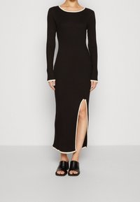 Robe en maille côtelée noire avec manches longues et col de couleur crème. Caractéristiques : fente latérale et coupe slim. Assortie à des sandales noires à enfiler.