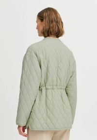 Chaqueta acolchada de color verde pastel con cintura ceñida por un cinturón, mangas amplias y un patrón de diamantes, con un cuello redondeado y sin cierres visibles.