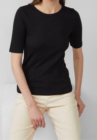 Svart ribbad kortärmad t-shirt med rund halsringning, figurnära passform, tillverkad av ett mjukt, stretchigt tyg. Bärs med ljusa byxor.