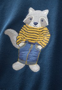 Broderi af en vaskebjørn iført en gulstribet sweater og blå jeans på en marineblå baggrund, med hvide poter og grå ansigtsdetaljer.