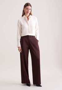 Crèmekleurige blouse met lange mouwen en een knoopsluiting, gecombineerd met bordeauxrode, hooggesneden broek met wijde pijpen. Gladde textuur, nauwsluitende pasvorm.