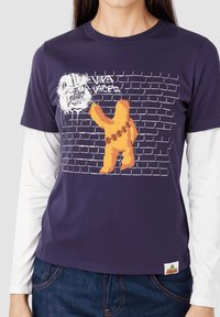 Camiseta de algodón púrpura con un gráfico de una figura naranja contra una pared de ladrillos blancos, con el texto "Viva Vaces" encima, en un estilo de arte urbano.