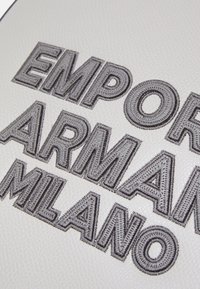 Relief ja tikandiga tekst "EMPORIO ARMANI MILANO" valgel tekstuurilisel nahast taustal.