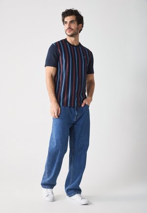 Tricou scurt în dungi, navy cu linii verticale roșii și albastre, purtat împreună cu blugi largi din denim albastru și adidași albi.