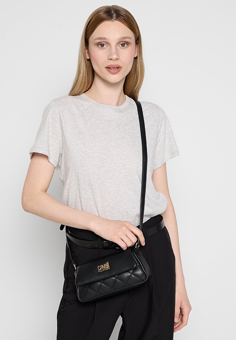 Giovane donna con capelli biondi lisci, che indossa una maglietta grigia chiara, pantaloni neri e tiene in mano una piccola borsa a tracolla nera trapuntata.