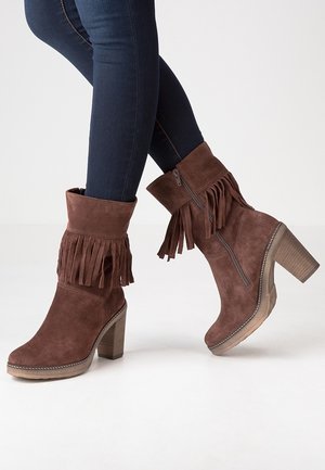 Bottes chevilles en daim marron avec franges, fermeture éclair latérale et talons empilés portées avec un jean skinny foncé sur un fond uni.