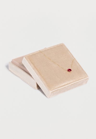 Caja de joyería de terciopelo beige con un delicado collar de cadena dorada que presenta un pequeño colgante en forma de corazón rojo, descansando sobre la tapa.