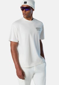 Hombre con camiseta blanca de North Sails, gorro blanco, gafas de sol deportivas con tintado púrpura y pantalones blancos, con una mano en el bolsillo.