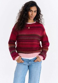 Sweter dziergany w różnych odcieniach czerwieni i różu, z okrągłym dekoltem, długimi rękawami oraz prążkowanym brzegiem, noszony na białej koszuli.