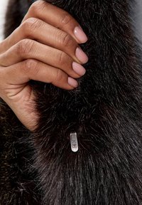 Sort faux fur beklædning med en blød tekstur. En hånd griber fat om kraven og afslører en sølv hardwaredetalje med præget mønster.