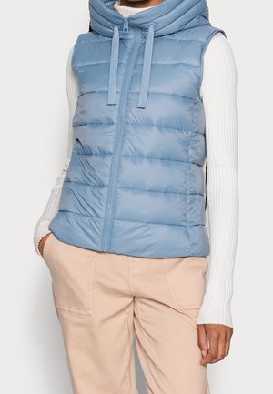Frau trägt eine hellblaue gesteppte Weste mit Kapuze über einem weißen gerippten Langarmshirt und beigen Hosen, Hände in den Taschen.