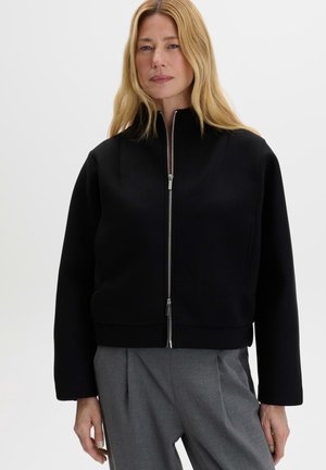 Donna con i capelli lunghi e biondi che indossa una giacca nera con zip e pantaloni grigi, in piedi davanti a uno sfondo bianco semplice.