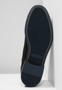 Semelle de sneaker noire avec surface en caoutchouc texturé, présentant un avant de semelle bleu prononcé et des coutures dorées le long du bord. Logo visible.