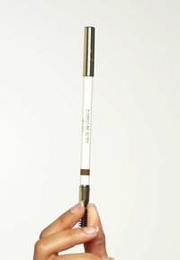 Matita per sopracciglia con corpo bianco e cappuccio dorato, caratterizzata da una tonalità marrone sulla punta e un pennello spoolie all'estremità opposta.