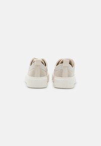 Beige joggesko med mesh-overdel, hvit gummisåle og bakre trekkstropp. Rund form og minimalistisk design med teksturerte detaljer.