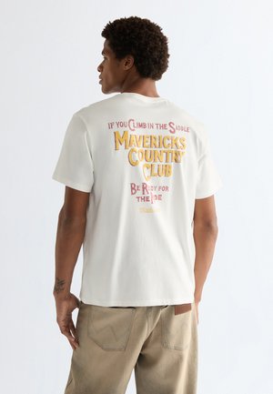 Jonge man met krullend haar, gekleed in een wit T-shirt met de tekst "Mavericks Country Club" en beige broek, staand tegen een witte achtergrond.