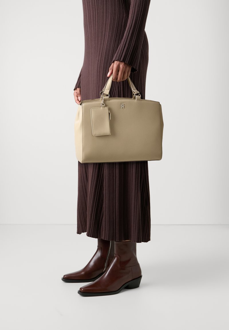Borsa strutturata beige con doppi manici, etichetta staccabile e accentuazione con logo. Tenuta da una persona che indossa un vestito lungo a coste marrone e stivali marroni.