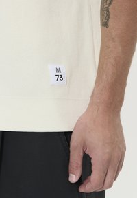 Světle krémové pletené tričko s malou bílou nášivkou s nápisem "M 73" v černé barvě. Tričko má texturovaný povrch a volný střih.