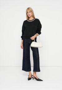 Sudadera negra con un escote adornado con joyas, combinada con jeans oscuros de pierna ancha y tacones negros de punta con tira al talón. Lleva un bolso blanco.