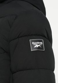 Czarna kurtka puchowa z pikowanym wzorem, miękką fakturą oraz naszywką z logo Reebok na rękawie.
